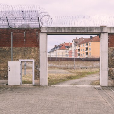 Ein Bild im Innenhof der JVA Rennelberg. Ein großes Tor in einer hohen Backsteinmauer, dahinter das ehemalige Gefängnisgelände. Über der Mauer verläuft Stacheldraht, im Hintergrund sind die umliegenden Wohnhäuser.